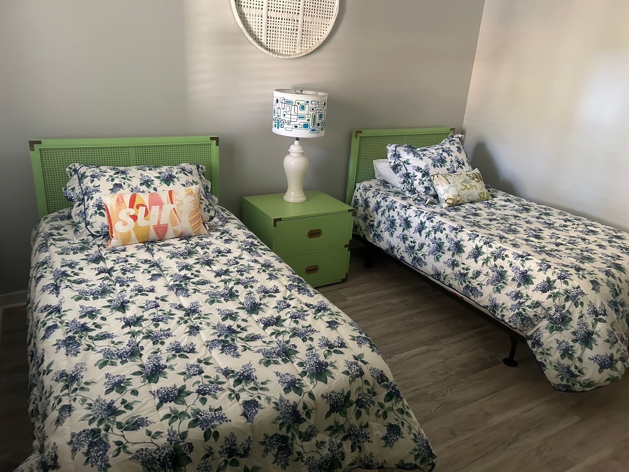 A bedroom with two single beds, each with a green headboard and floral bedding. There is a nightstand with a lamp between the beds.