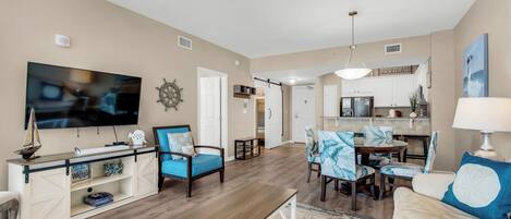 A spacious living room with a TV, seating, a dining area, and a kitchen in the background.