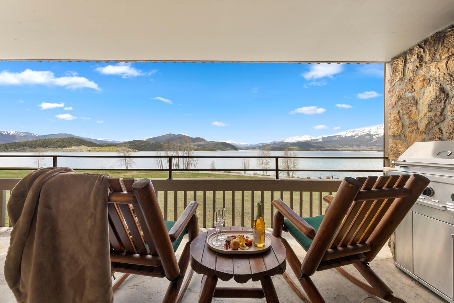 A balcony with two wooden chairs, a small round table with a tray of food and drinks, and a grill. A mountain and lake are visible in the background.