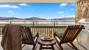Un balcón con dos sillas de madera, una mesita redonda con una bandeja de comida y bebidas, y una parrilla. Se ven una montaña y un lago al fondo.