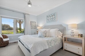 A bedroom with a large bed, a nightstand with a lamp, a chair, and a window with a view of palm trees. - NEW PHOTOS! Stylish, Fully Renovated Sea Dunes Condo on Ground Floor! (Fernandina Beach)