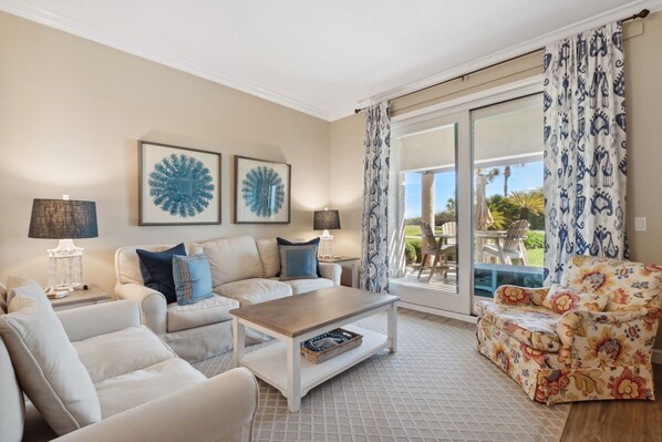 A living room with a sofa, armchairs, a coffee table, and a patterned armchair. There are two wall art pieces, a lamp, and a window with patterned curtains. - NEW PHOTOS! Stylish, Fully Renovated Sea Dunes Condo on Ground Floor! (Fernandina Beach)