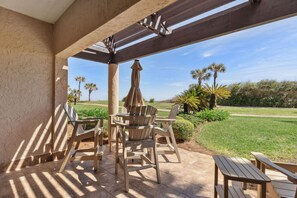 An outdoor patio with wooden chairs and tables, a pergola, and a view of a well-maintained lawn and palm trees. - NEW PHOTOS! Stylish, Fully Renovated Sea Dunes Condo on Ground Floor! (Fernandina Beach)
