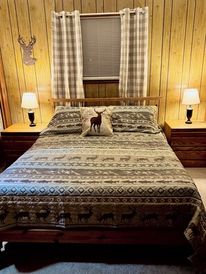Una cama con una colcha estampada, dos lámparas de noche, un cabecero de madera y una ventana con persianas.