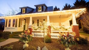 Una casa de dos pisos con un porche decorado con luces y coronas, un jardín delantero con plantas en macetas y un cielo despejado.
