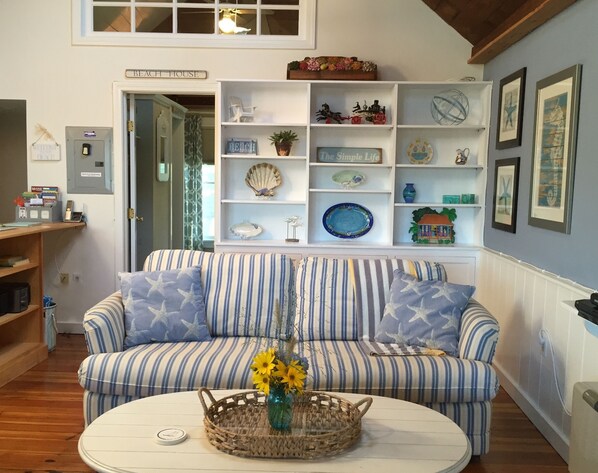 A room with a striped sofa, a round coffee table with a vase of flowers, a white shelving unit with decorative items, and a sign reading "BEACH HOUSE".