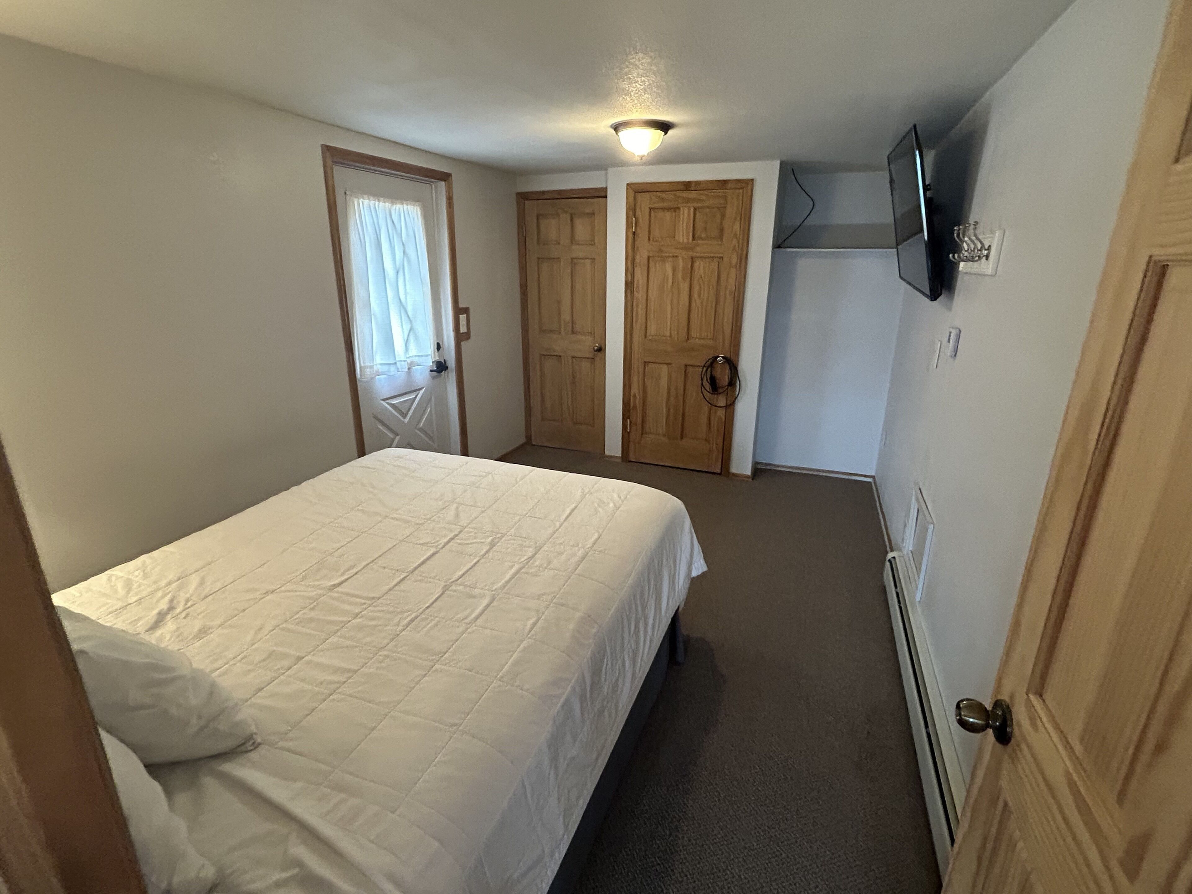A bedroom with a bed, two wooden doors, a ceiling light, and a wall-mounted TV.