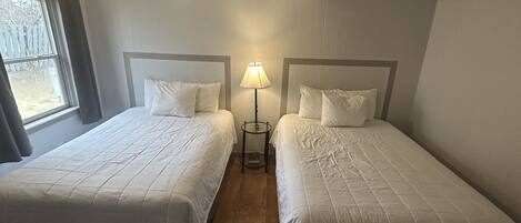 A room with two single beds, each with a headboard, a bedside table with a lamp, and a window with curtains.