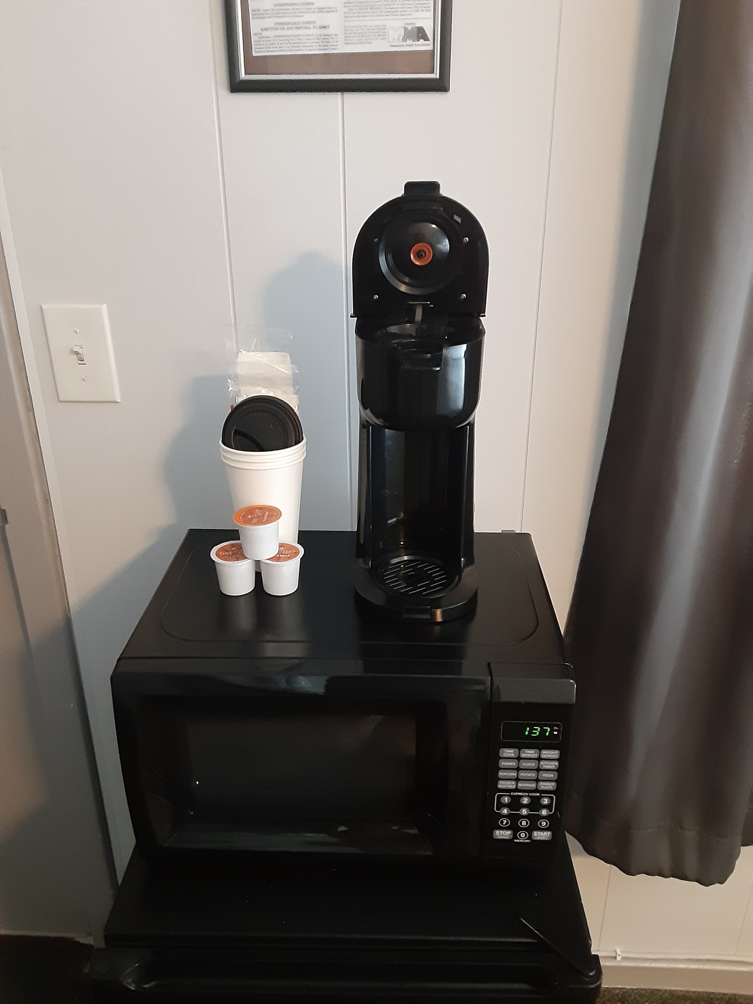 Una cafetera negra sobre una encimera negra, con pantalla digital, dos vasos desechables blancos y un vaso blanco con tapa.