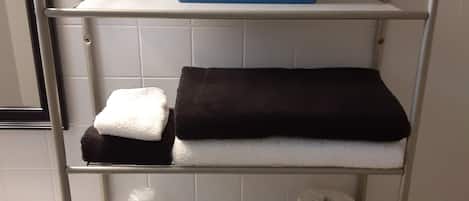 Een badkamer met toilet, wastafel en een metalen plank waarop handdoeken, toiletpapier en een pak tissues liggen.