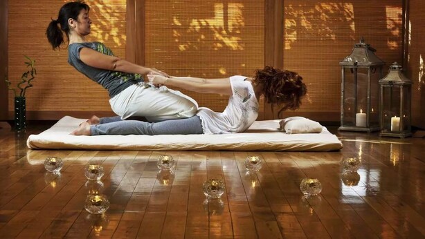 Eine Person führt eine Yoga-Pose auf einer Matte in einem Raum mit Holzboden, einem Fenster mit Jalousien und zwei beleuchteten Laternen auf einem Tisch aus.