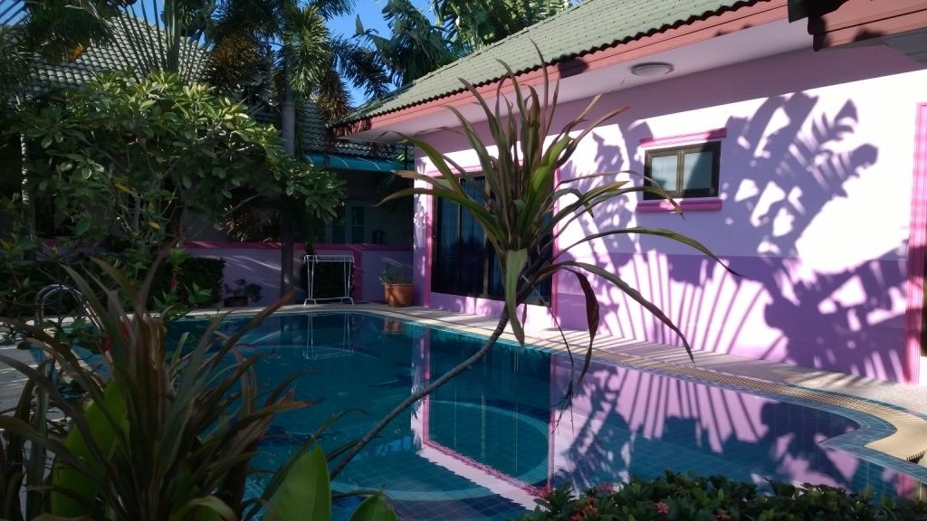 A swimming pool surrounded by lush greenery and a pink building with a tiled roof.