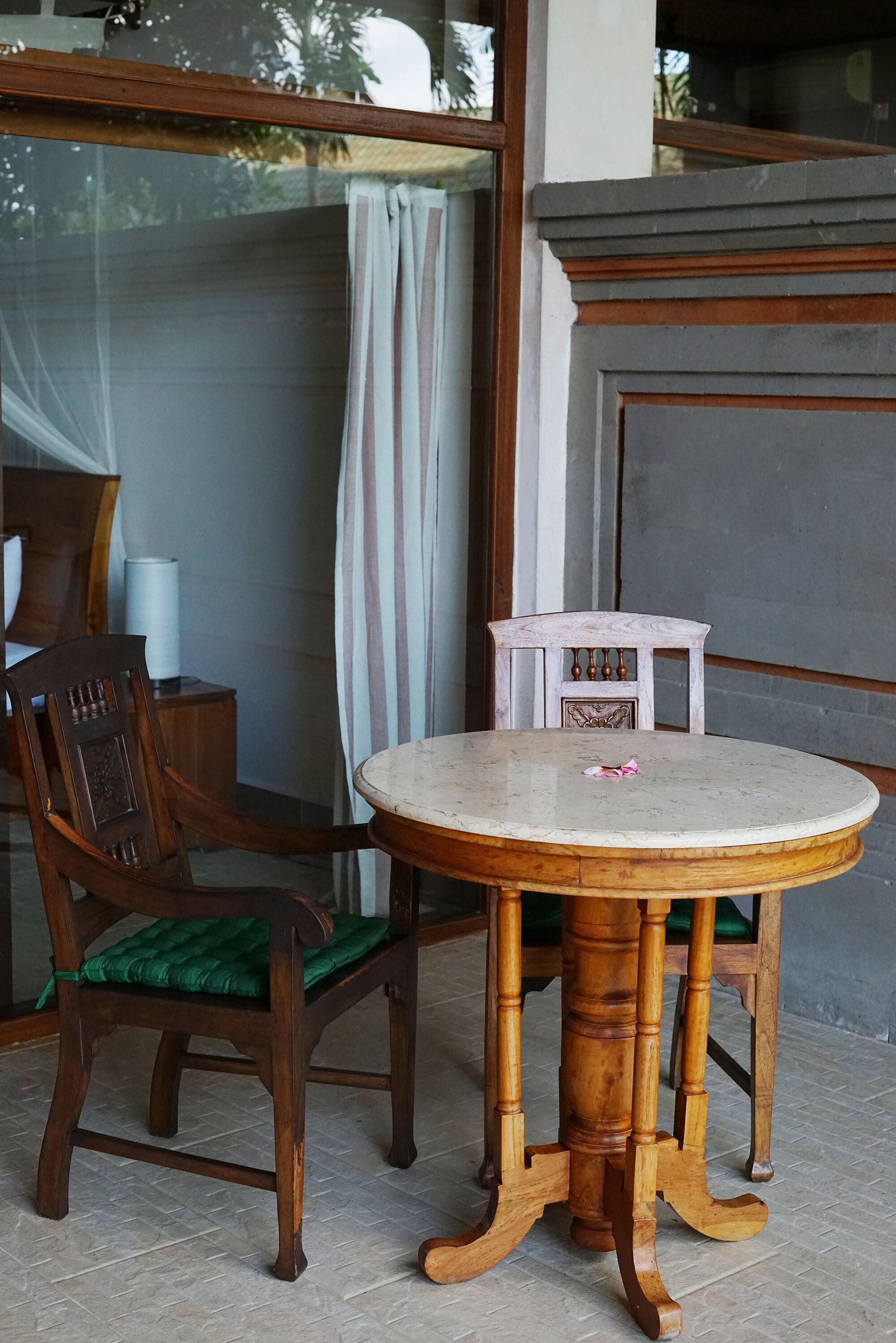 A round wooden table with a green cushioned chair, a small white cabinet, and a window with a curtain.