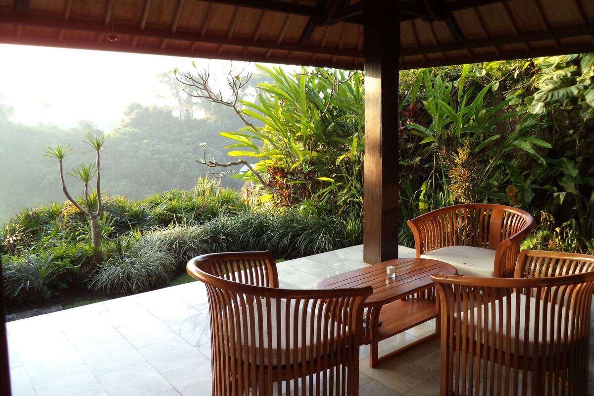 A wooden table and chairs under a roofed patio with a view of lush greenery and trees.