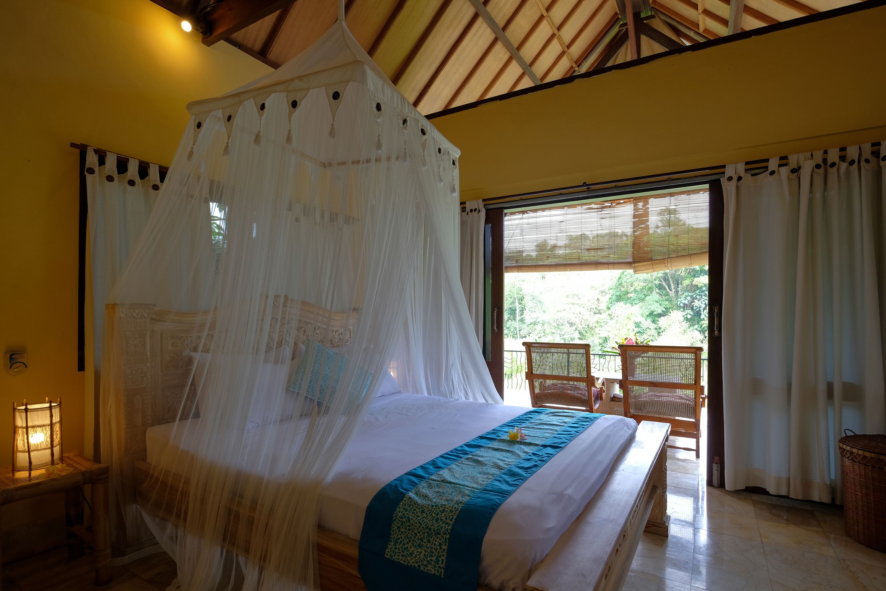 Una cama con dosel, una mesita de noche con lámpara y vistas al exterior a través de una ventana.