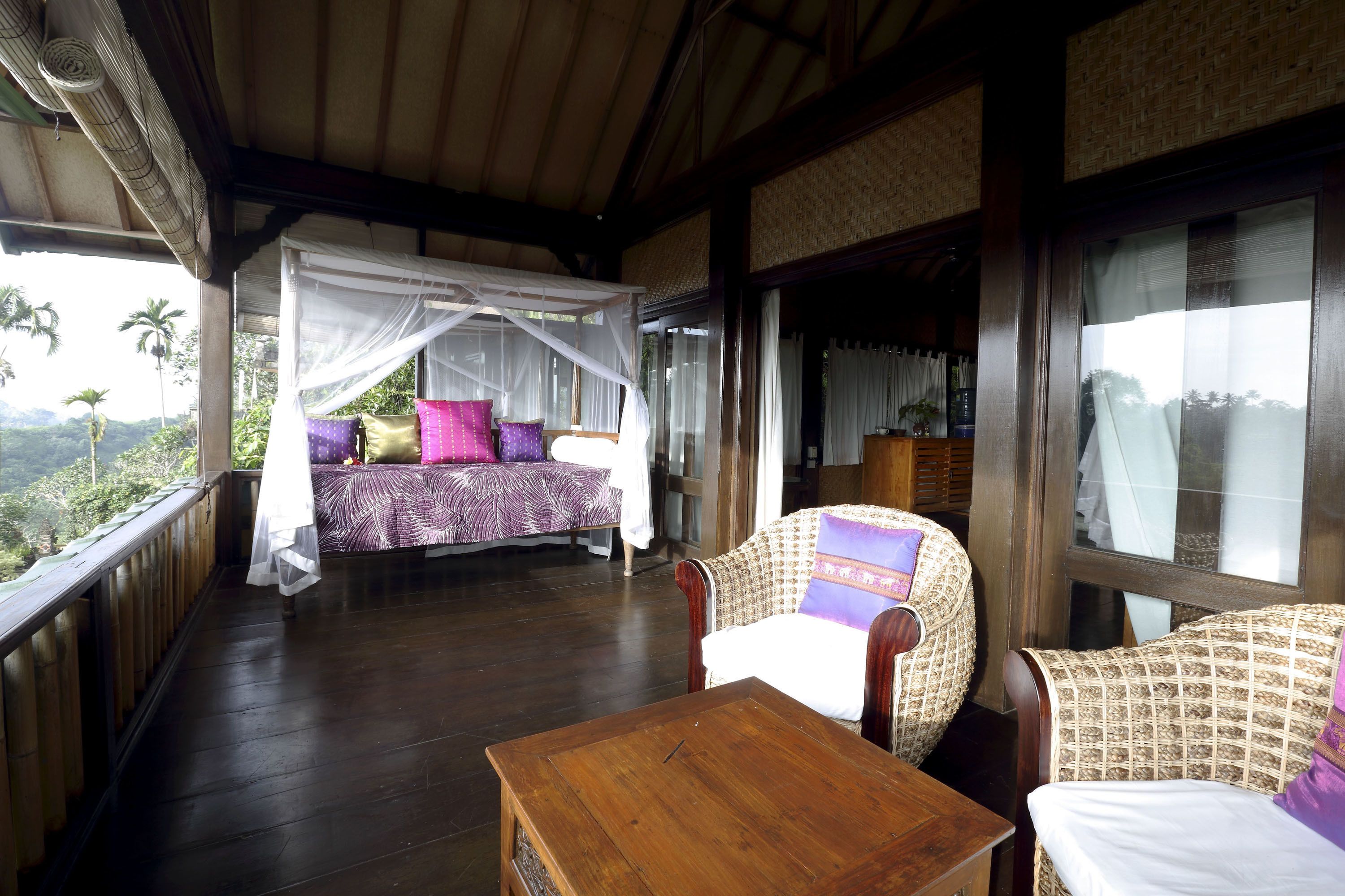 A room with a bed under a mosquito net, a wooden table, and a view of the outdoors.