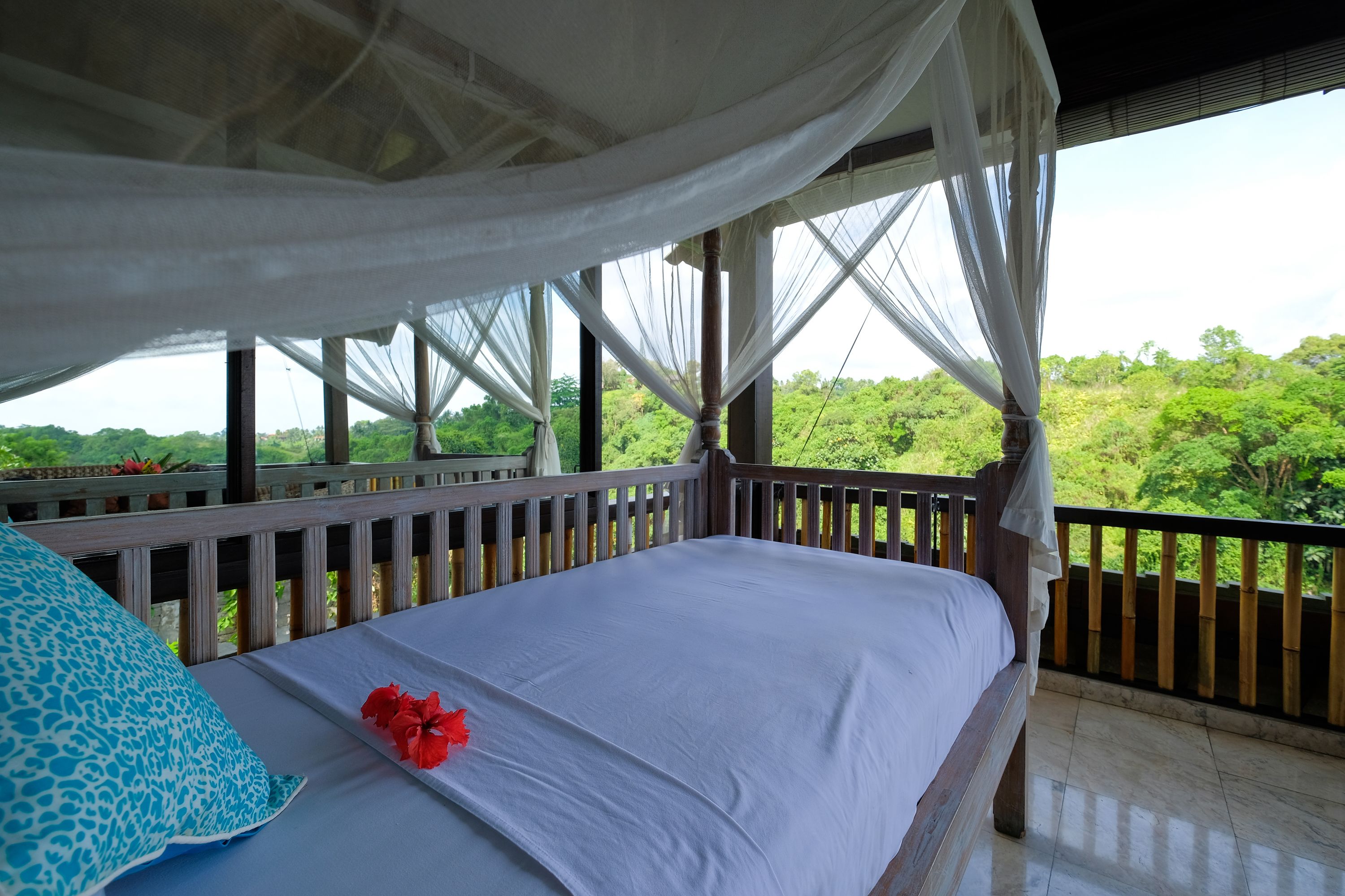 A bed with a white sheet and a blue pillow, a red flower, and a wooden railing.