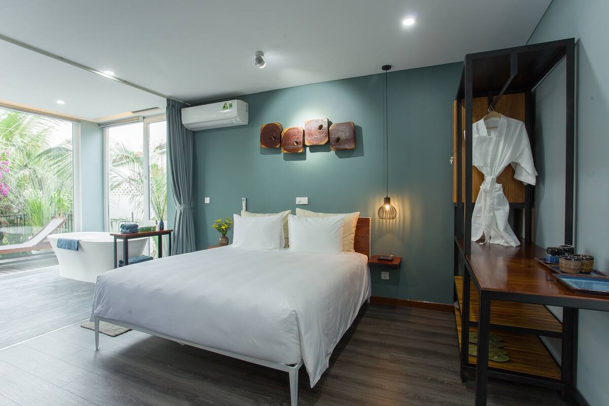 A modern bedroom with a large bed, a bedside table, a mirror, and a view of the outdoors.