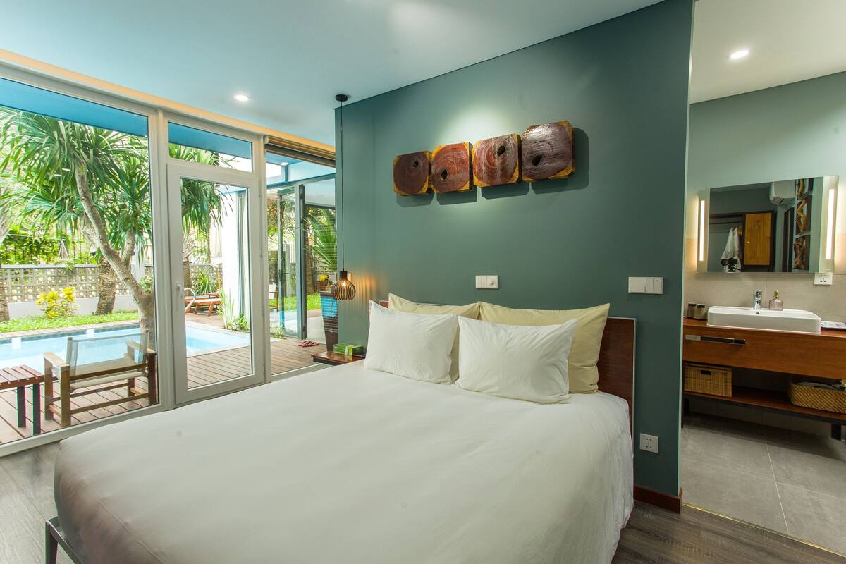 A modern bedroom with a large bed, a bathroom with a sink and mirror, and a view of a swimming pool.