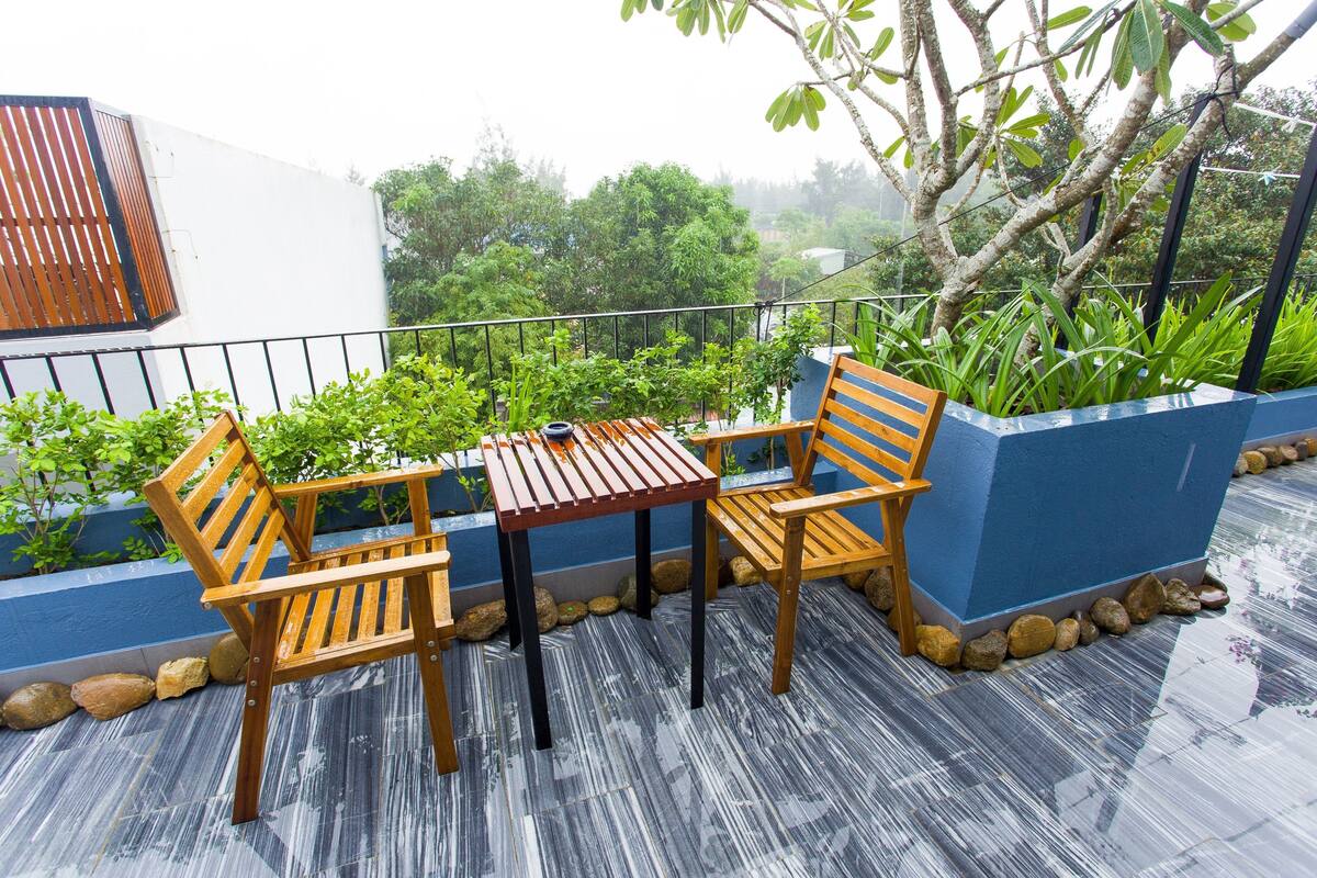 A wooden table and chairs on a balcony with a view of greenery and a building.