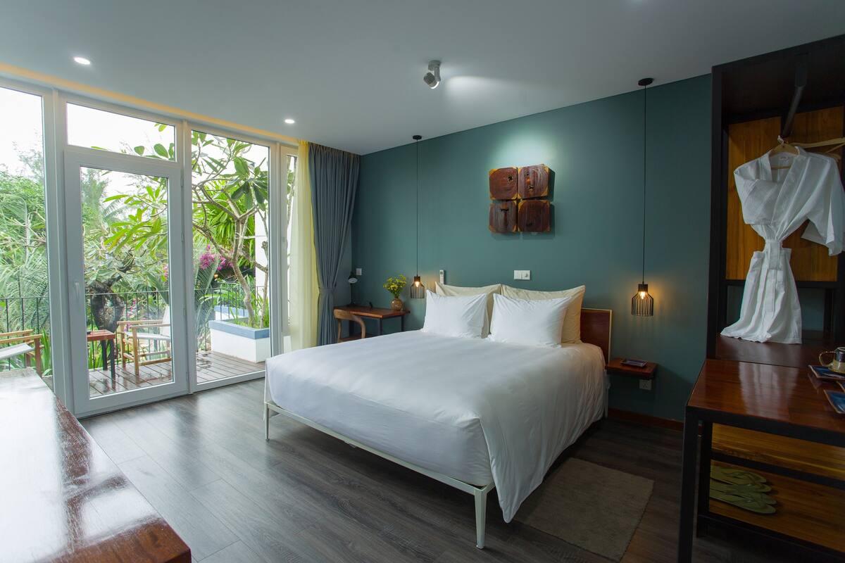 A bedroom with a large bed, a wooden desk, a nightstand, and a wardrobe. There is a sliding glass door leading to a balcony with plants.