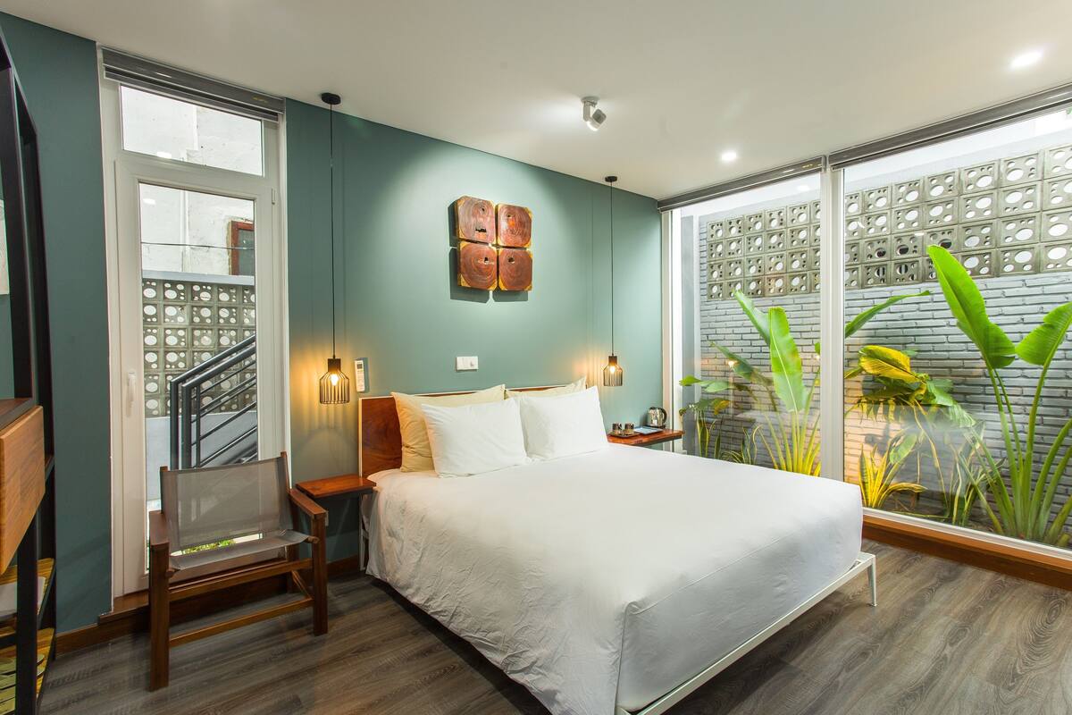 A bedroom with a bed, a bench, a nightstand, and a window with a view of plants.