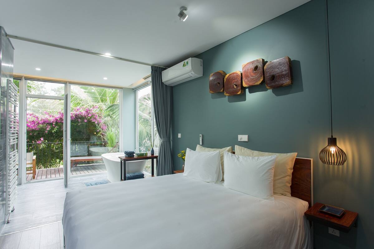 A modern bedroom with a large bed, a desk, and a view of a balcony with plants.