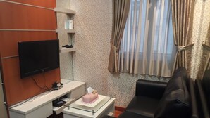 Een kleine, moderne kamer met een flatscreen televisie aan de muur, een wit bureau met een computer en een teddybeer, een zwarte leren bank en een raam met gordijnen.