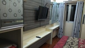Een slaapkamer met een televisie aan een muur, een bed met een decoratieve spreideken, een bureau met een stoel, en een raam met gordijnen.