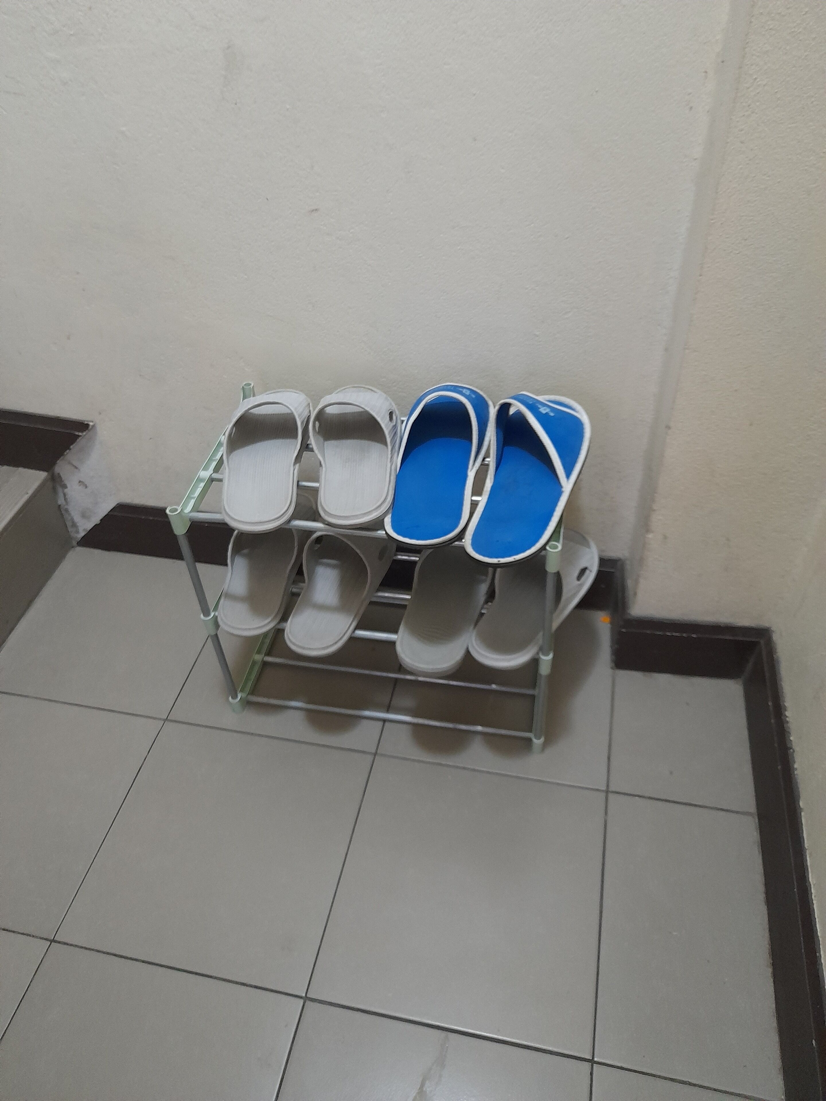 Un estante metálico con varios pares de zapatillas, algunas con el interior azul, frente a una pared con suelo de baldosas.