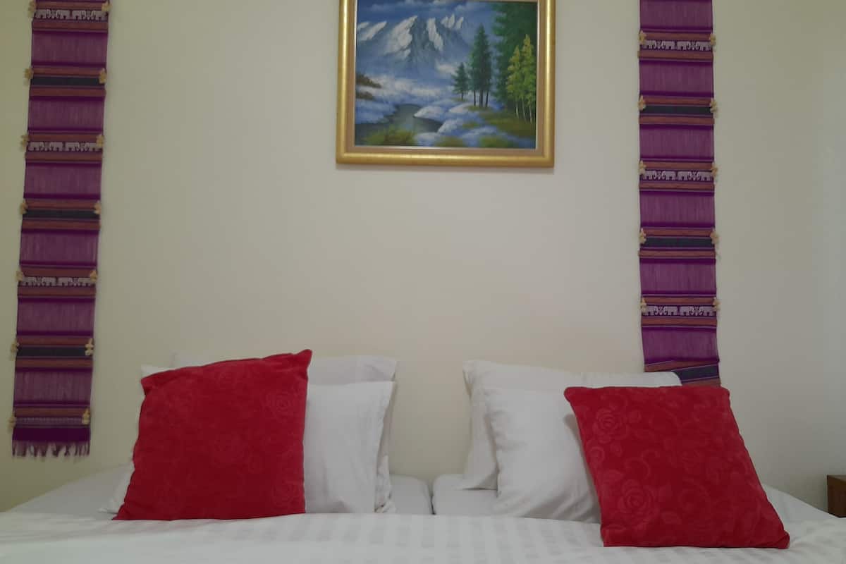 A bed with white and red pillows, a painting of a mountain landscape, and purple patterned drapes on the walls.