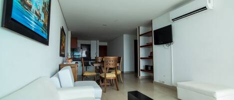 Una sala de estar moderna con un sofá blanco, un comedor con sillas de madera, un televisor de pantalla plana montado en la pared y un equipo de aire acondicionado.