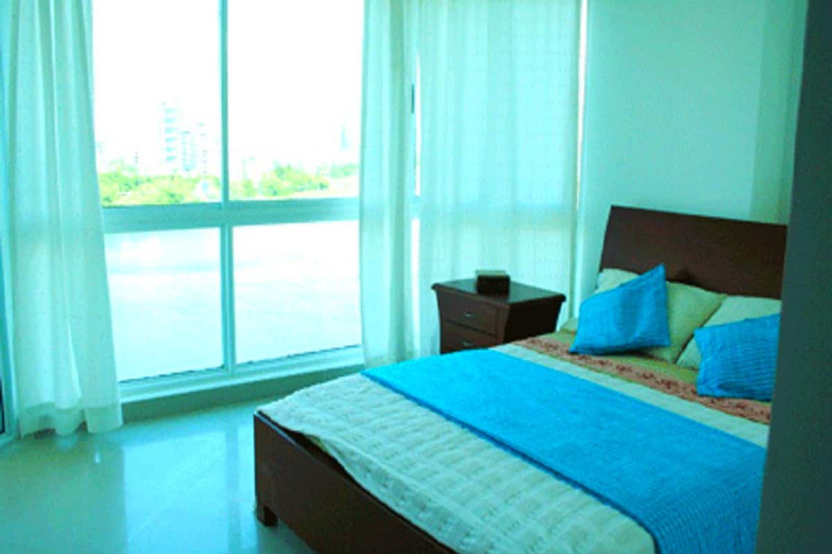 A bedroom with a large window, a bed with blue bedding, and a nightstand.