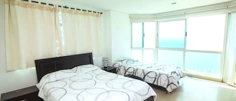 A bedroom with a large bed, a smaller bed, a nightstand, and a window with a view of the sea.