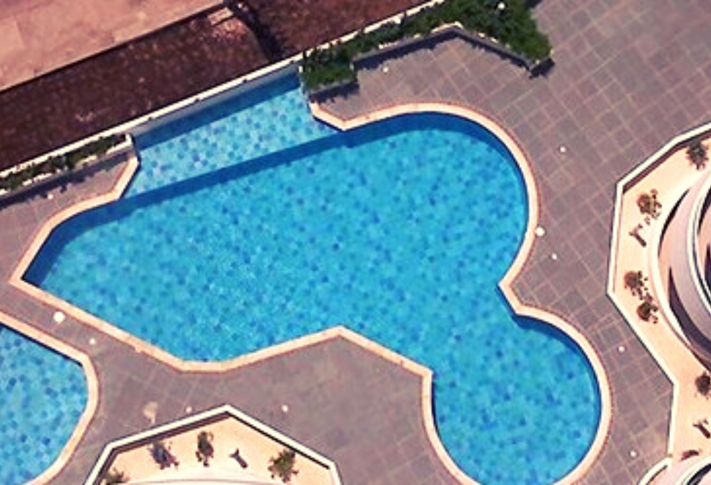 An aerial view of a swimming pool with a unique geometric shape, surrounded by a paved area with a low wall and a small garden.