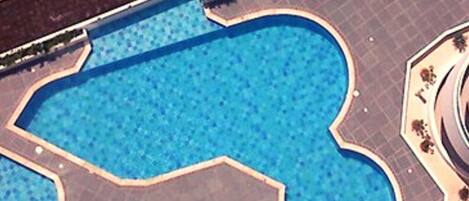 An aerial view of a swimming pool with a unique geometric shape, surrounded by a paved area with a low wall and a small garden.
