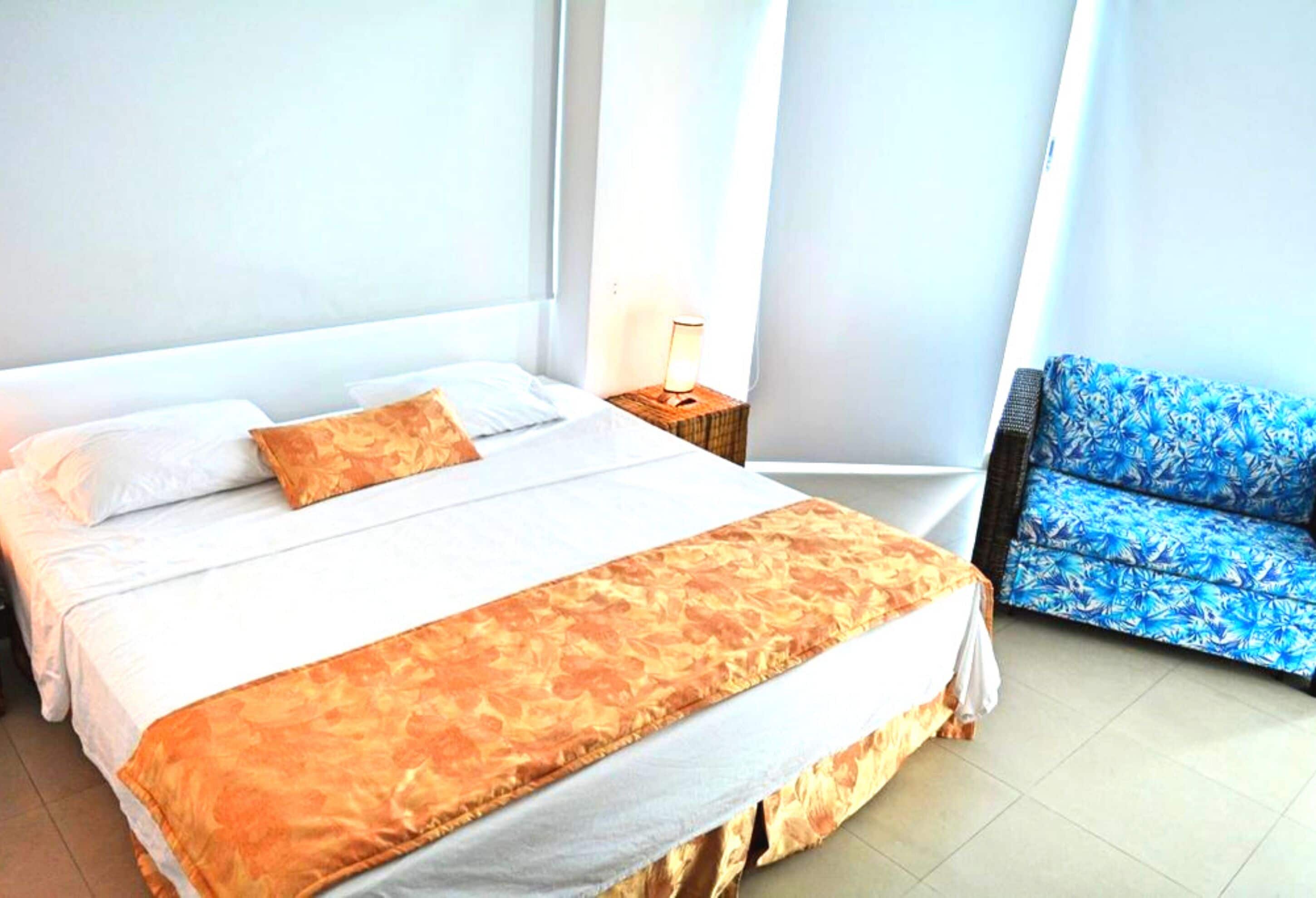 A hotel room with a bed, a nightstand, a lamp, and a blue patterned chair.