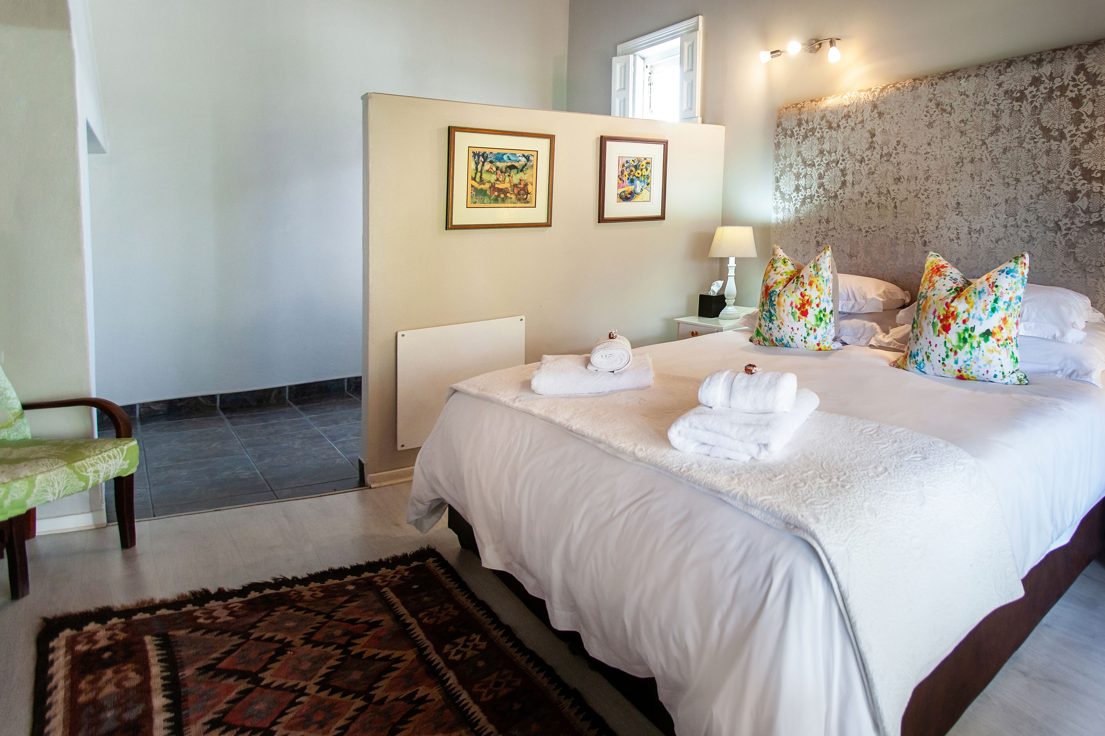 Une chambre d’hôtel avec un grand lit, une table de chevet avec une lampe, deux œuvres d’art encadrées au mur et un tapis à motifs.