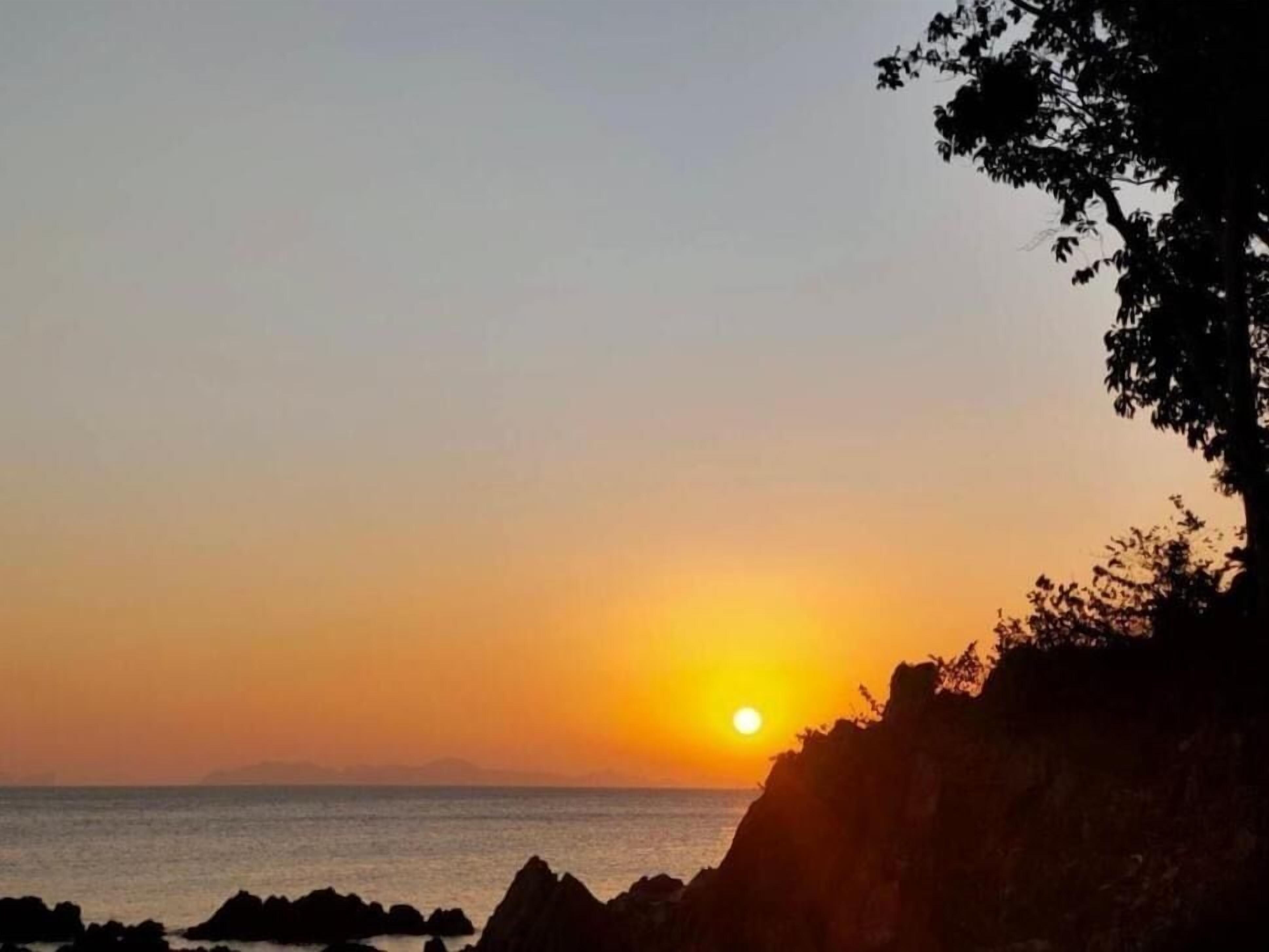 A sunset over a coastal area with rocky formations and a tree line.