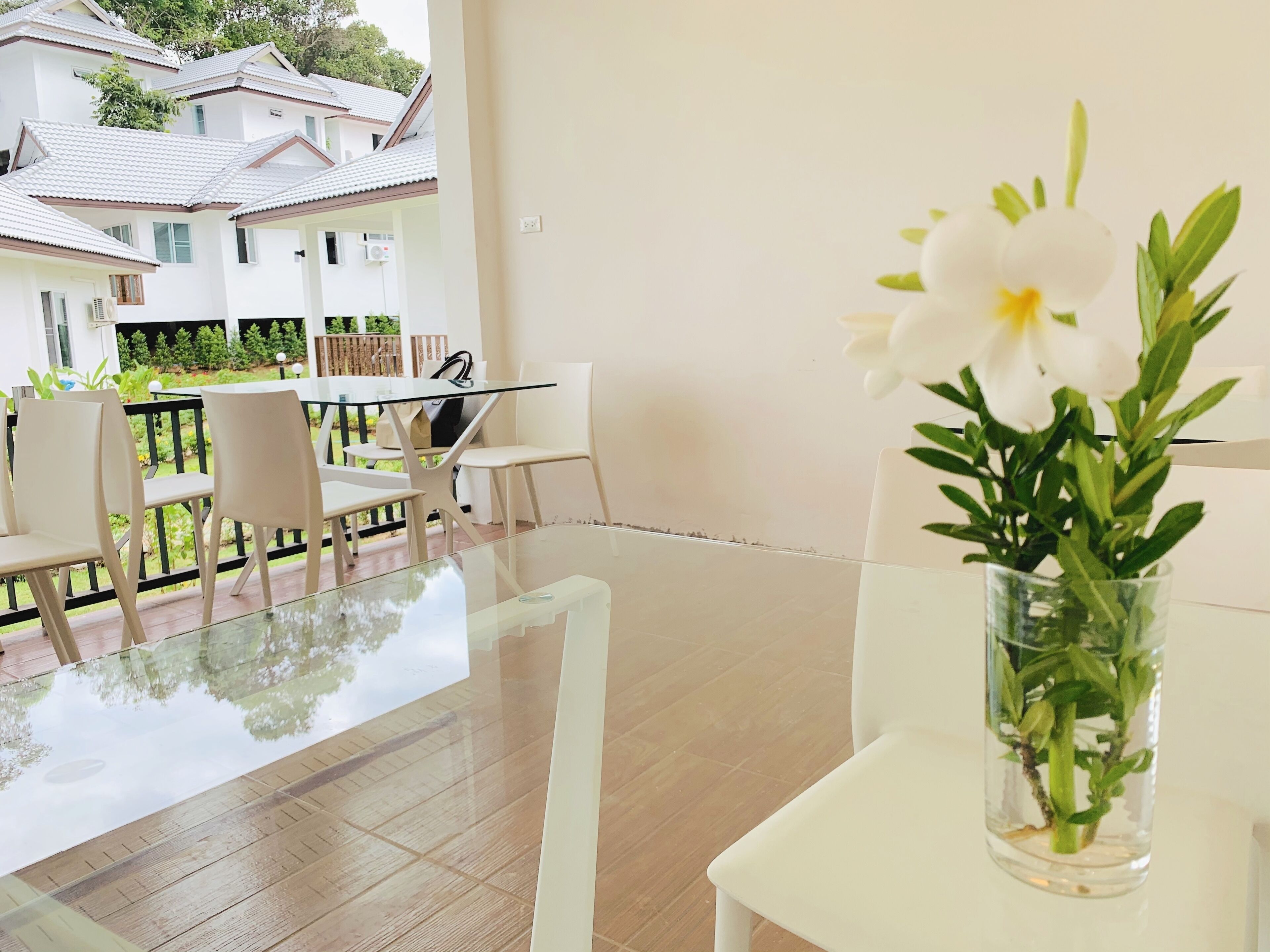 A modern room with a glass table, white chairs, a vase with white flowers, and a view of a white building with a tiled roof.