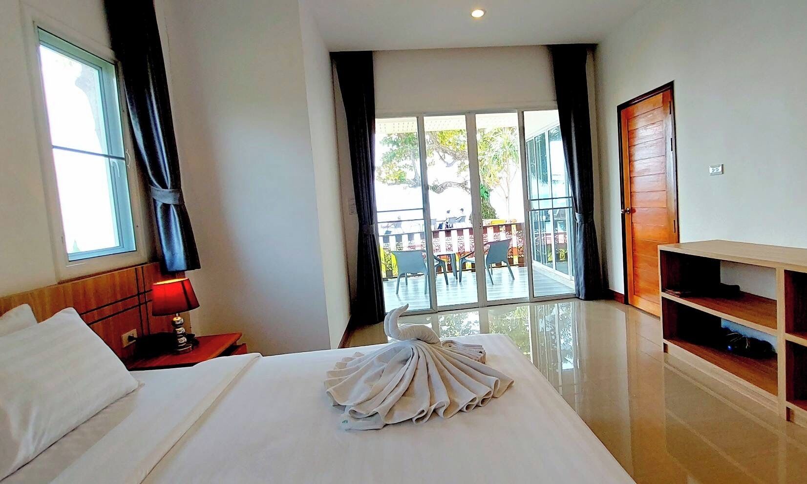 Habitación de hotel con una cama grande, una mesita de noche con lámpara, un mueble de televisión de madera y un balcón con mobiliario exterior.