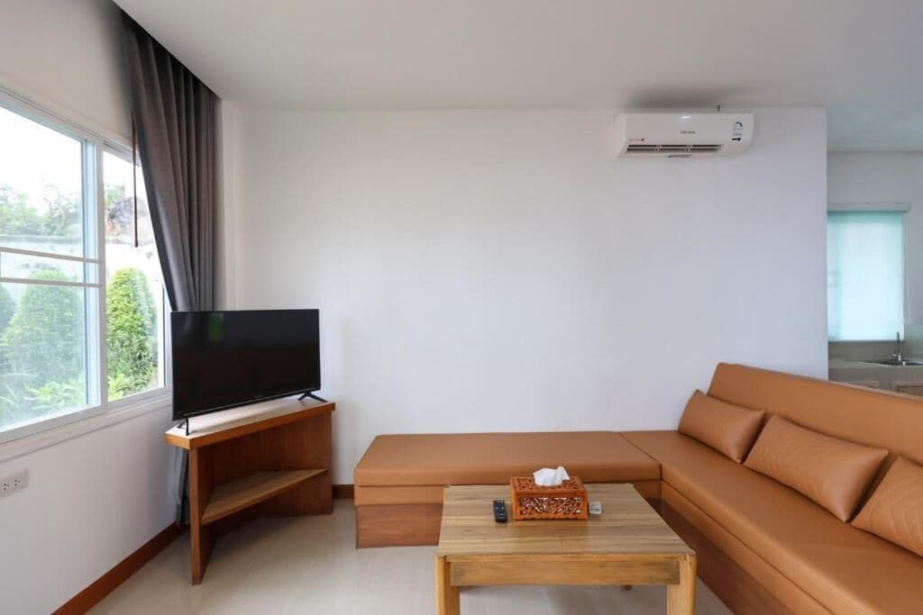 A modern living room with a flat-screen TV on a wooden stand, a brown leather sofa, a wooden coffee table, and a large window with curtains.