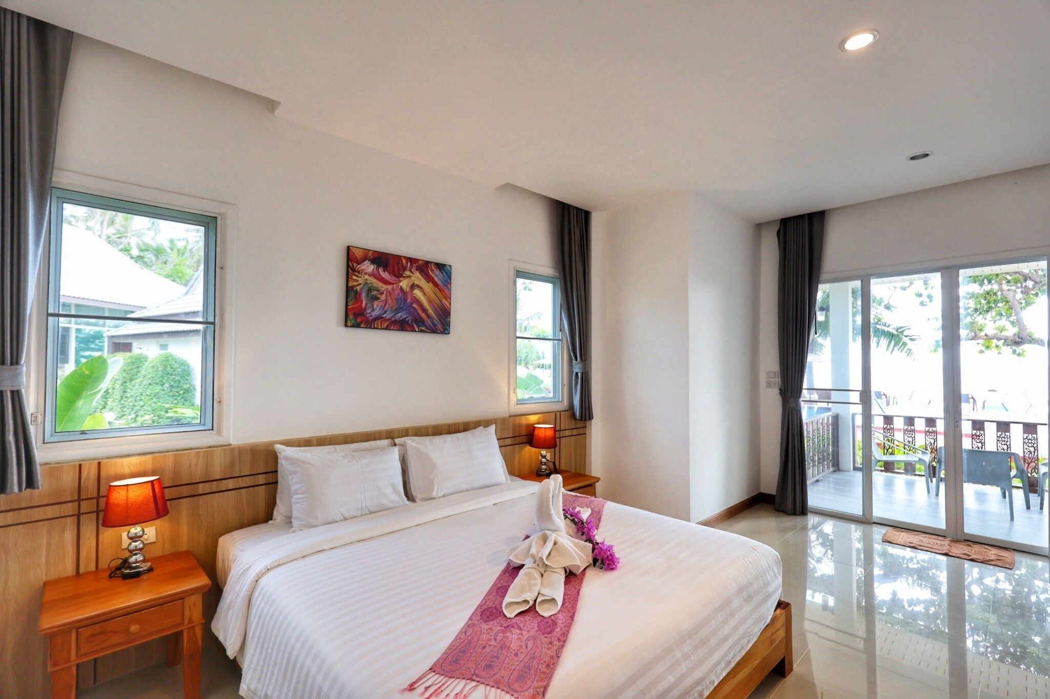 A hotel room with a large bed, a nightstand with a lamp, a painting on the wall, and a view of the outdoors through the window.