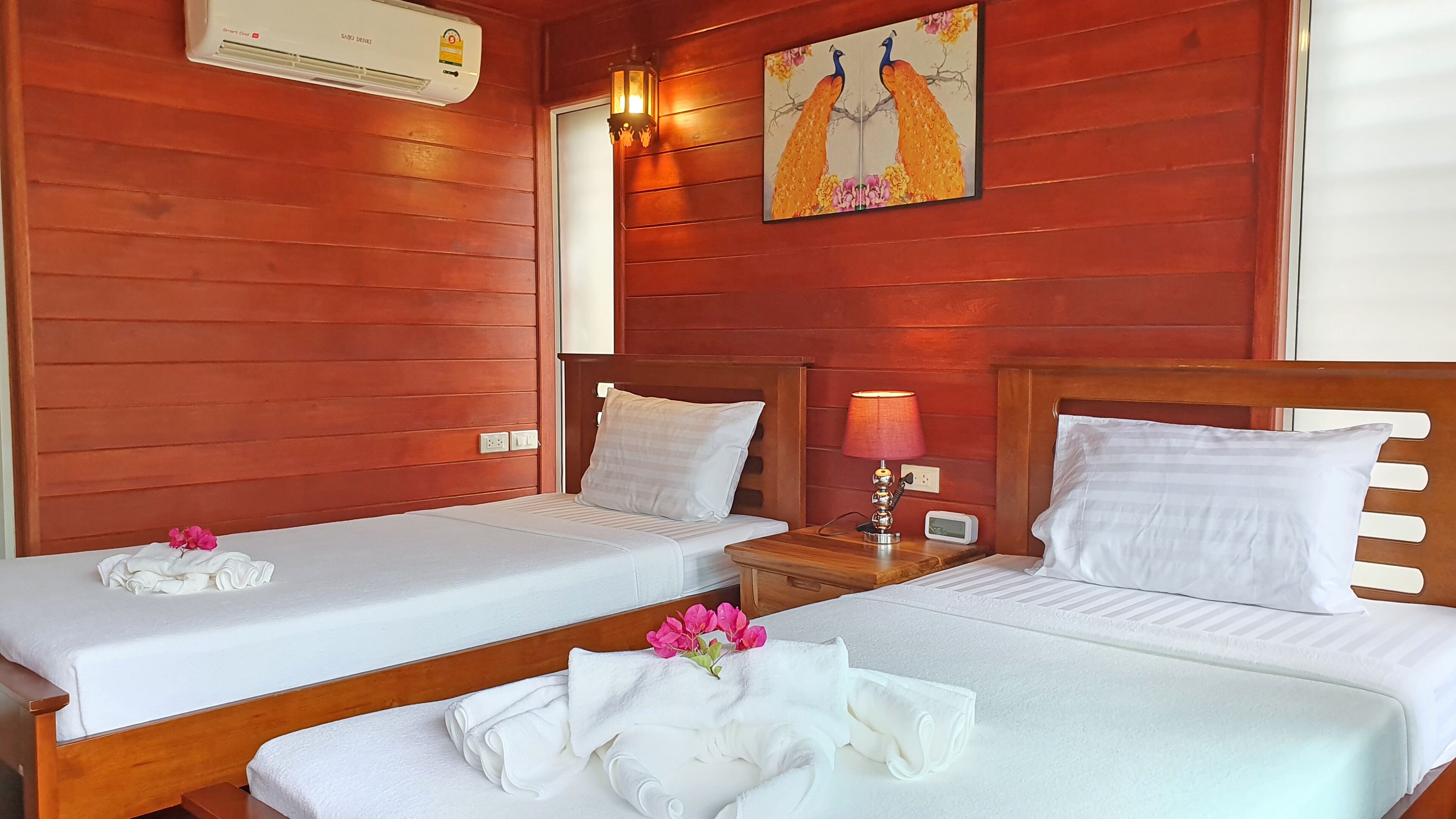 A hotel room with two single beds, wooden headboards, white linens, a wall-mounted air conditioner, a nightstand with a lamp, and a painting of a peacock.