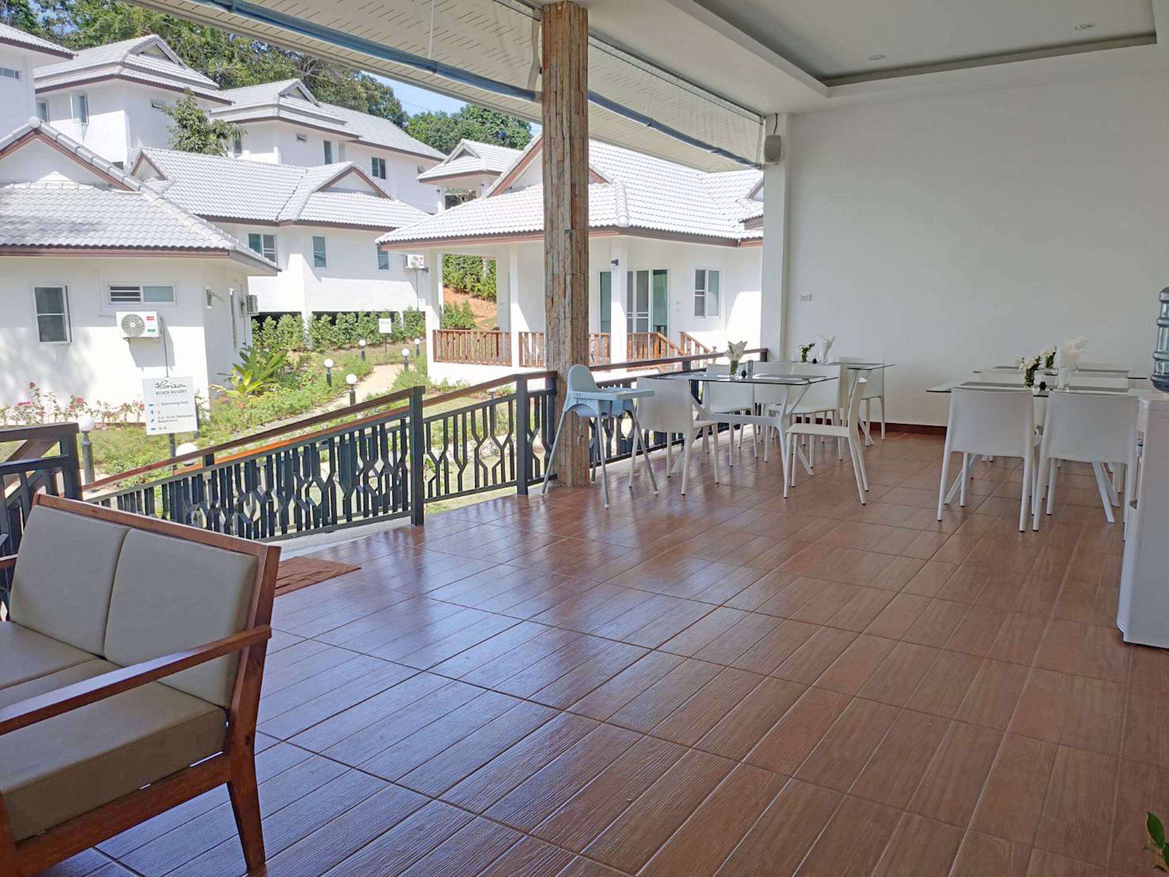 Un balcón amplio con piso de madera, mobiliario blanco y vista a edificios blancos al fondo.