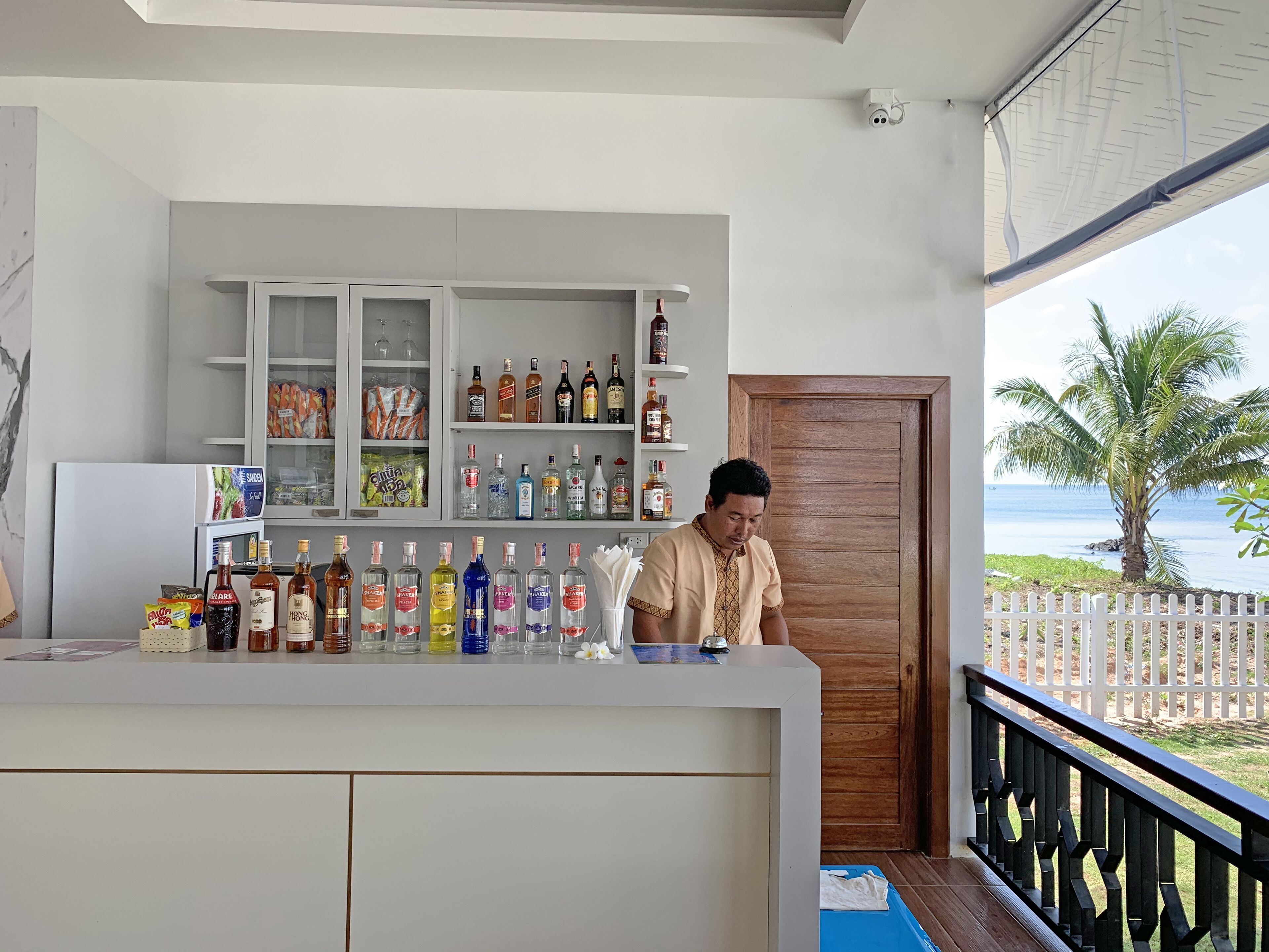 A bar area with a variety of liquor bottles, a refrigerator, and a wooden door leading to a balcony with a view of the sea.