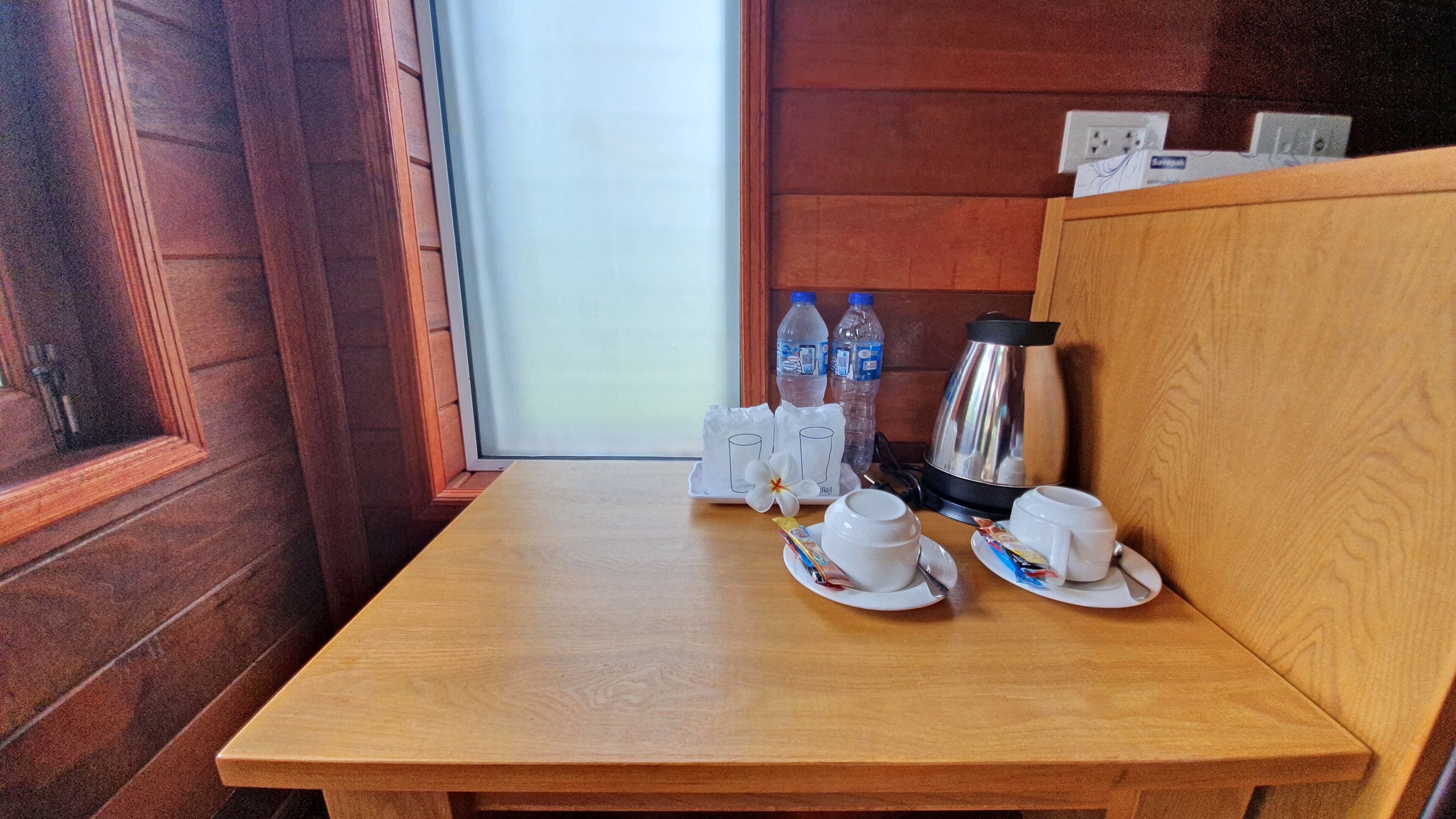 Une table en bois avec deux tasses et sous-tasses blanches, une fleur et une cafetière.