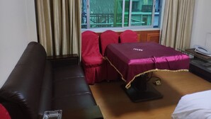 Una habitación con un sofá de cuero marrón oscuro, una mesita con un mantel rojo, una cama con colcha roja y una ventana con cortinas.