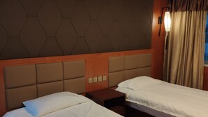 Una habitación de hotel con dos camas individuales, un cabecero con diseño geométrico, una mesita de noche con lámpara y una ventana con cortinas.