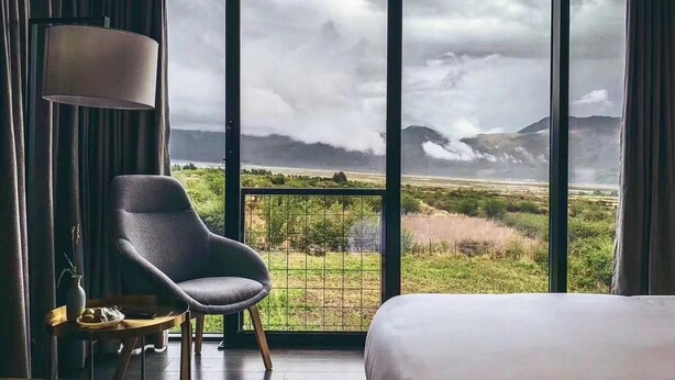 A room with a bed, a chair, and a large window offering a view of a landscape with mountains and a cloudy sky. - The Rock Hotel (Lijiang)